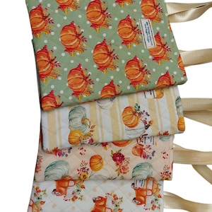 Może przedstawiać: Stos pikowanych przedmiotów z tkaniny z motywami jesiennymi. Górny element przedstawia pomarańczowe dynie na jasnym zielonym tle. Inne przedmioty przedstawiają dynie, kwiaty i motywy jesienne. Dołączone są beżowe wstążki.