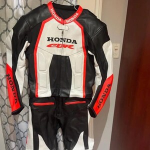 Könnte beinhalten: Schwarzer, weißer und roter Honda CBR Motorrad-Rennanzug. Der Anzug ist aus Leder gefertigt und trägt das Honda-Logo auf Brust, Schultern und Armen. Der Anzug hängt an einem Kleiderbügel.