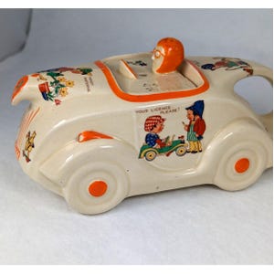 May include: A cream-colored ceramic teapot shaped like a vintage car with orange accents. It features illustrations of children, including one labeled "YOUR LICENCE PLEASE!" and another "WENDY WATERING". The lid has an orange driver figure.