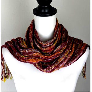 May include: A multi-colored scarf with a fringed edge is draped over a white mannequin. The scarf features a blend of burgundy, orange, yellow, and purple hues. The texture appears to be woven, with a slightly shiny finish.