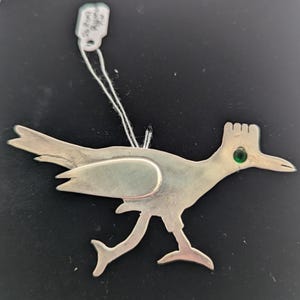 May include: A silver-toned metal roadrunner ornament with a green gem eye. The bird is depicted in a running pose, with its tail feathers spread. A tag is attached to the top with a string.