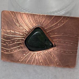 May include: A copper-colored rectangular brooch with a dark green, triangular stone at its center. The brooch has an embossed sunburst design with curved lines and dotted accents. The metal has a textured, hammered appearance.