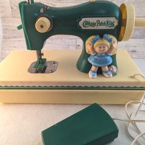 May include: A green and cream-colored Cabbage Patch Kids toy sewing machine with a matching green foot pedal. The machine features a molded figure of a blonde doll in a blue dress on the side and includes a white power cord and a hand wheel.