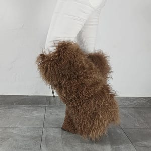 May include: Tall brown boots featuring long, curly, shaggy fur texture covering the entire exterior. The boots reach mid-calf and are worn with white denim pants and a plaid shirt. The design includes a thin cord detail visible on the side.