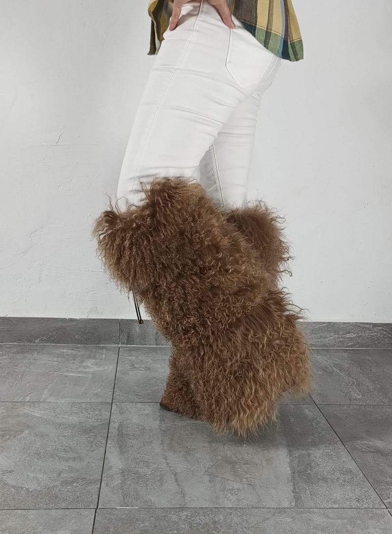 May include: Tall brown boots featuring long, curly, shaggy fur texture covering the entire exterior. The boots reach mid-calf and are worn with white denim pants and a plaid shirt. The design includes a thin cord detail visible on the side.