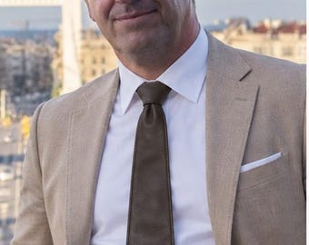 Peut inclure: Un blazer beige porté sur une chemise blanche à col boutonné avec une cravate en cuir marron foncé. Le blazer présente une pochette blanche dans la poche de poitrine, et une ceinture en cuir marron est visible à la taille du pantalon assorti.
