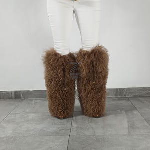 May include: A pair of tall, brown, curly-textured fur boot covers worn over white pants. The boot covers feature a prominent black logo with the letter Z and the word FURS positioned between the two legs, with small gold-toned hardware accents.