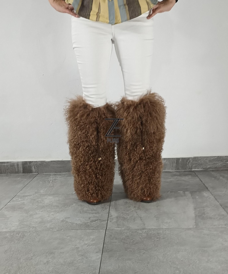 May include: A pair of tall, brown, curly-textured fur boot covers worn over white pants. The boot covers feature a prominent black logo with the letter Z and the word FURS positioned between the two legs, with small gold-toned hardware accents.