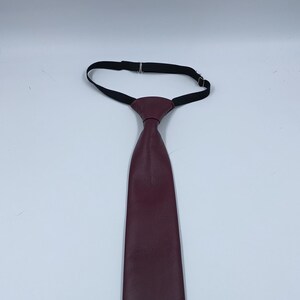 Peut inclure: Une cravate bordeaux avec une texture lisse rappelant le cuir et une extr&eacute;mit&eacute; pointue. La cravate pr&eacute;sente un n&oelig;ud pr&eacute;-nou&eacute; et une sangle de cou &eacute;lastique noire r&eacute;glable avec une boucle en m&eacute;tal pour la fixer autour du cou.