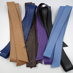 Könnte beinhalten: Eine Auswahl an Ledergürteln in verschiedenen Farben, darunter Beige, Marineblau, Braun, Lila, Schwarz und Hellblau. Die Gürtel sind lang und schmal, mit einem spitz zulaufenden Ende. Das Bild ist gut beleuchtet und zeigt die Textur und Farbunterschiede.