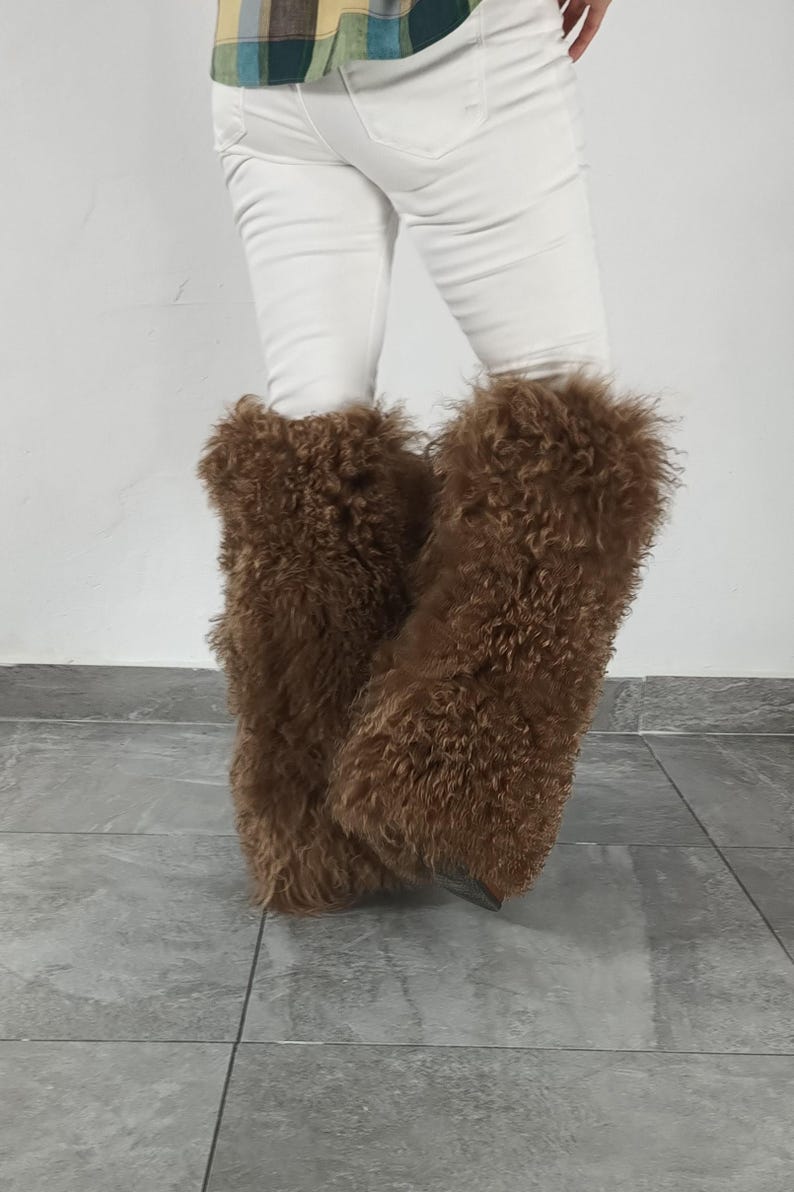 May include: A pair of brown, long-haired, shaggy faux fur leg warmers worn over white trousers. The leg warmers feature a dense, textured, curly pile that extends from the lower calf down to the ankle, providing a voluminous and fuzzy appearance.