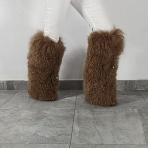 May include: A pair of tall, brown, shaggy faux fur leg warmers or boot covers with a textured, curly appearance. The material is thick and voluminous, extending from the lower calf to the ankle, worn over white trousers against a plain wall.