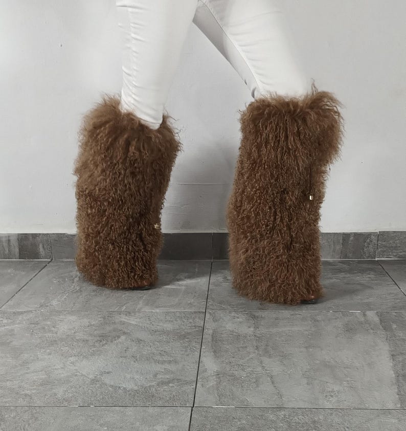 May include: A pair of tall, brown, shaggy faux fur leg warmers or boot covers with a textured, curly appearance. The material is thick and voluminous, extending from the lower calf to the ankle, worn over white trousers against a plain wall.