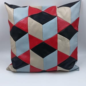 May include: Square decorative pillow with a geometric pattern. The pillow features a patchwork design with red, black, beige, and light blue leather pieces. The pillow is on a white background.