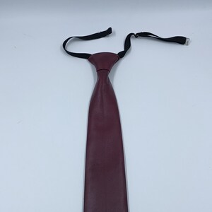 Peut inclure: Une cravate bordeaux fonc&eacute; avec une texture lisse rappelant le cuir. La cravate pr&eacute;sente un n&oelig;ud pr&eacute;-nou&eacute; et une sangle de cou &eacute;lastique noire r&eacute;glable avec un crochet et un m&eacute;canisme coulissant en m&eacute;tal argent&eacute; pour fixer l'accessoire.