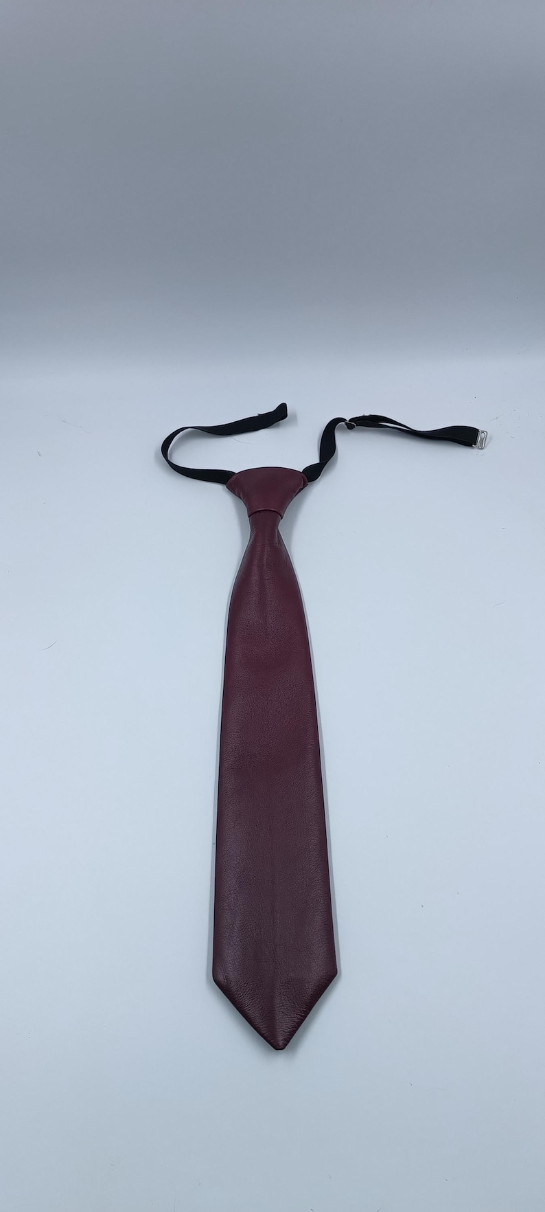 Peut inclure: Une cravate bordeaux fonc&eacute; avec une texture lisse rappelant le cuir. La cravate pr&eacute;sente un n&oelig;ud pr&eacute;-nou&eacute; et une sangle de cou &eacute;lastique noire r&eacute;glable avec un crochet et un m&eacute;canisme coulissant en m&eacute;tal argent&eacute; pour fixer l'accessoire.