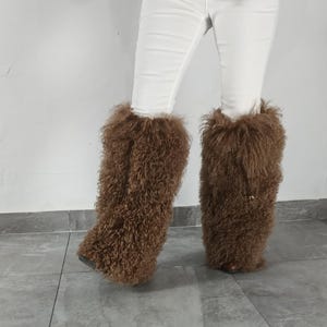 May include: A pair of brown, long, curly faux fur leg warmers worn over white pants. The leg warmers feature a thick, shaggy texture that covers the lower legs, extending from the mid-calf down to the feet, with a visible drawstring detail.