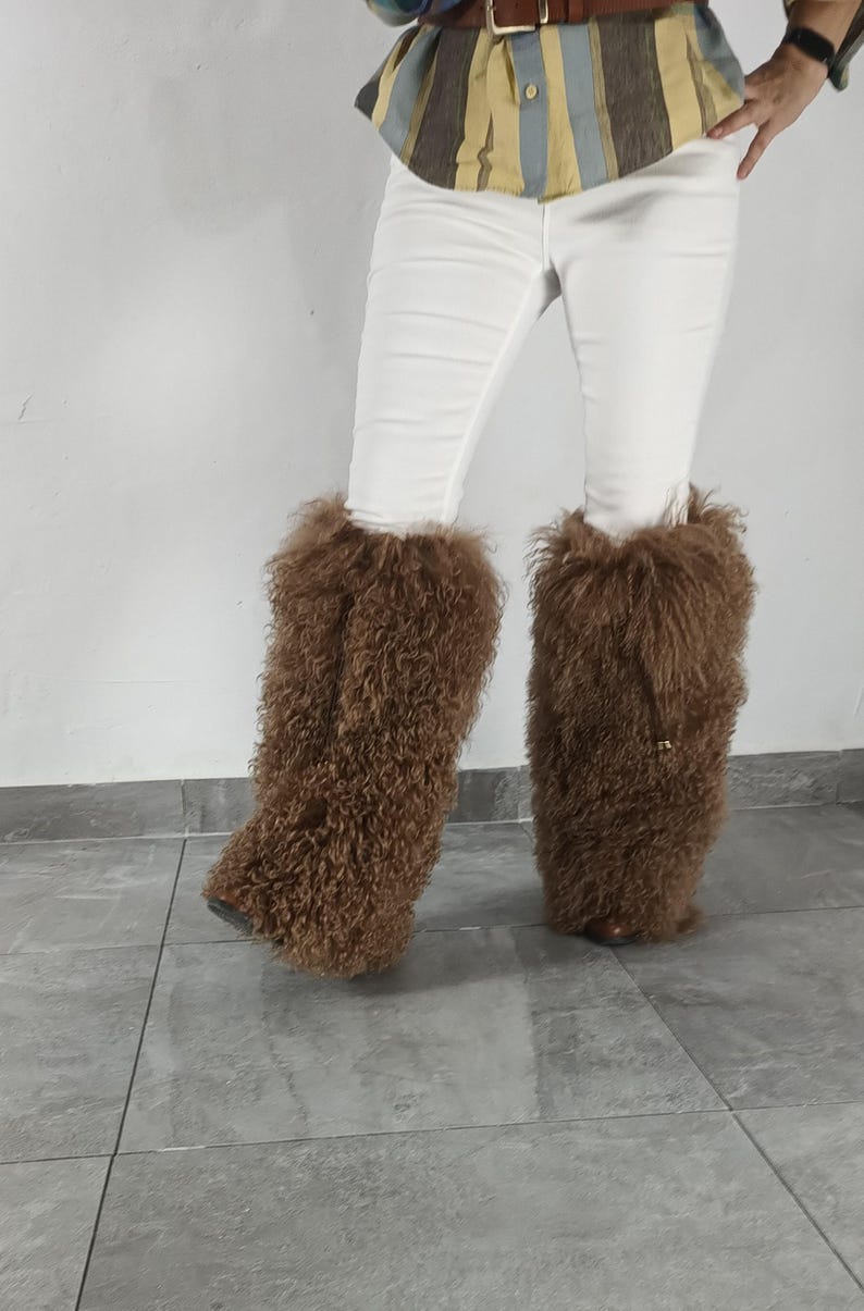 May include: A pair of brown, long, curly faux fur leg warmers worn over white pants. The leg warmers feature a thick, shaggy texture that covers the lower legs, extending from the mid-calf down to the feet, with a visible drawstring detail.