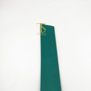 Peut inclure: Une longue bande &eacute;troite de mat&eacute;riau vert fonc&eacute; uni. La surface est lisse et pr&eacute;sente une texture mate. Pr&egrave;s du haut de la bande se trouve un logo de couleur dor&eacute;e repr&eacute;sentant les lettres stylis&eacute;es TS avec le mot FURS imprim&eacute; en dessous.