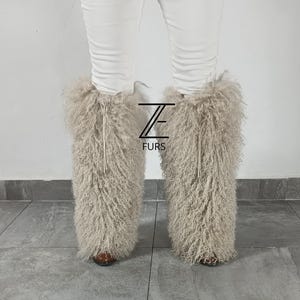 May include: A pair of beige, long-haired, curly fur leg warmers worn over white pants. The leg warmers feature a drawstring closure at the top and a logo with the text ZE FURS positioned between the two legs. The texture is shaggy and dense.