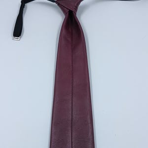 Peut inclure: Une cravate en cuir bordeaux profond pr&eacute;sentant une surface lisse et textur&eacute;e avec une couture verticale centrale sur toute la longueur. La cravate comprend une sangle noire r&eacute;glable avec un curseur en m&eacute;tal argent&eacute; pour le cou.