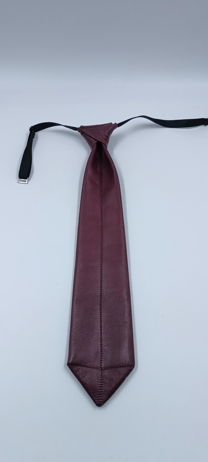 Peut inclure: Une cravate en cuir bordeaux profond pr&eacute;sentant une surface lisse et textur&eacute;e avec une couture verticale centrale sur toute la longueur. La cravate comprend une sangle noire r&eacute;glable avec un curseur en m&eacute;tal argent&eacute; pour le cou.