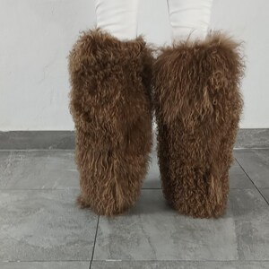 May include: A pair of brown, shaggy, long-haired faux fur leg warmers worn over white pants. The leg warmers feature a dense, curly, and textured appearance that covers the lower legs, providing a voluminous and fuzzy look from the knee down.