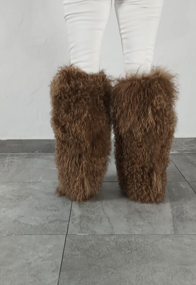 May include: A pair of brown, shaggy, long-haired faux fur leg warmers worn over white pants. The leg warmers feature a dense, curly, and textured appearance that covers the lower legs, providing a voluminous and fuzzy look from the knee down.