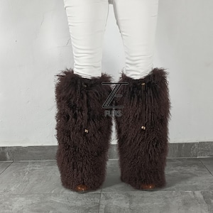 May include: A pair of dark brown, long-haired fur leg warmers worn over white pants. The leg warmers feature a drawstring closure at the top with gold-toned hardware. A logo with the text ZE FURS is visible in the center between the two legs.