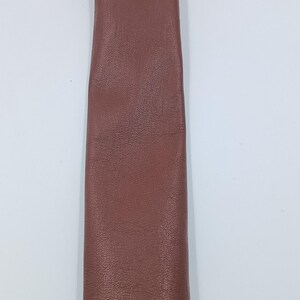 Peut inclure: Une longue bande &eacute;troite de cuir marron avec une texture grain&eacute;e visible. Le mat&eacute;riau est lisse et d'une couleur uniforme, pr&eacute;sentant une finition mate sur toute sa surface. La bande est positionn&eacute;e verticalement sur un fond blanc uni.