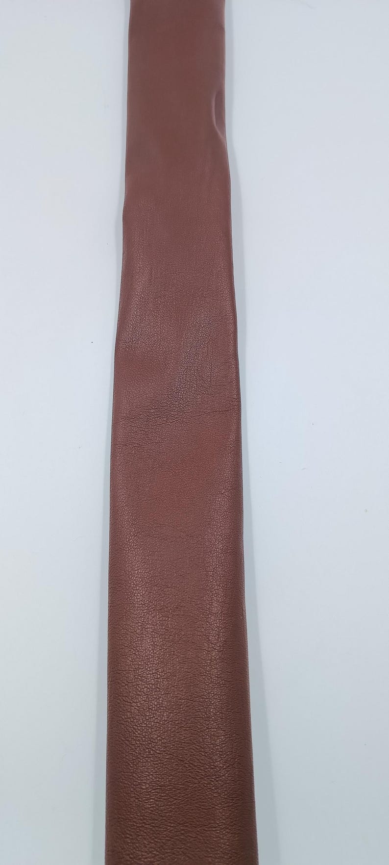 Peut inclure: Une longue bande &eacute;troite de cuir marron avec une texture grain&eacute;e visible. Le mat&eacute;riau est lisse et d'une couleur uniforme, pr&eacute;sentant une finition mate sur toute sa surface. La bande est positionn&eacute;e verticalement sur un fond blanc uni.