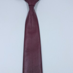 Peut inclure: Une cravate en cuir bordeaux profond pr&eacute;sentant une surface lisse et textur&eacute;e avec une extr&eacute;mit&eacute; pointue. La cravate est fix&eacute;e &agrave; une fine sangle de cou &eacute;lastique noire r&eacute;glable avec une fermeture &agrave; crochet en m&eacute;tal pour un ajustement facile.