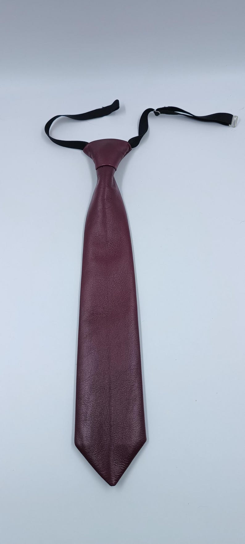 Peut inclure: Une cravate en cuir bordeaux profond pr&eacute;sentant une surface lisse et textur&eacute;e avec une extr&eacute;mit&eacute; pointue. La cravate est fix&eacute;e &agrave; une fine sangle de cou &eacute;lastique noire r&eacute;glable avec une fermeture &agrave; crochet en m&eacute;tal pour un ajustement facile.