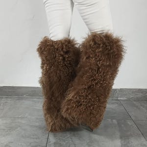May include: A pair of brown, long-haired, shaggy faux fur leg warmers worn over white trousers. The leg warmers feature a dense, textured, curly pile that extends from the lower calf down to the ankle, providing a voluminous and fuzzy appearance.