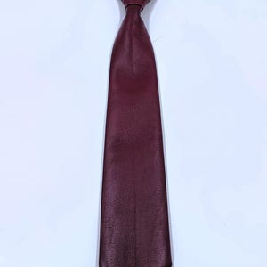 Peut inclure: Une cravate pr&eacute;-nou&eacute;e en cuir bordeaux profond &agrave; la surface textur&eacute;e. La cravate pr&eacute;sente une extr&eacute;mit&eacute; pointue et est fix&eacute;e &agrave; une fine sangle de cou &eacute;lastique noire r&eacute;glable, dot&eacute;e d'une boucle m&eacute;tallique pour l'ajuster autour du col.