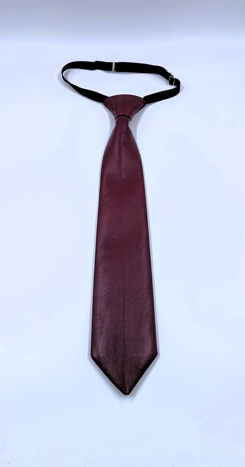 Peut inclure: Une cravate pr&eacute;-nou&eacute;e en cuir bordeaux profond &agrave; la surface textur&eacute;e. La cravate pr&eacute;sente une extr&eacute;mit&eacute; pointue et est fix&eacute;e &agrave; une fine sangle de cou &eacute;lastique noire r&eacute;glable, dot&eacute;e d'une boucle m&eacute;tallique pour l'ajuster autour du col.
