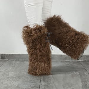 May include: A pair of brown, long-haired, fluffy fur leg warmers worn over white pants. The leg warmers feature a textured, curly appearance and include a small black logo patch with the text Z FURS visible on the front of the left leg warmer.