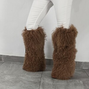 May include: A pair of brown, knee-high boots featuring a long, shaggy, curly fur texture that covers the entire shaft. The boots are worn with white trousers, and the design includes a small, dark vertical strap detail on the outer side.