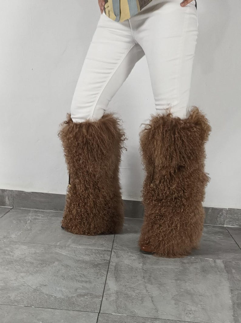 May include: A pair of brown, knee-high boots featuring a long, shaggy, curly fur texture that covers the entire shaft. The boots are worn with white trousers, and the design includes a small, dark vertical strap detail on the outer side.