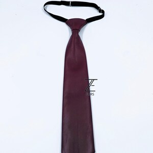 Peut inclure: Une cravate bordeaux fonc&eacute; avec une surface textur&eacute;e et une extr&eacute;mit&eacute; pointue. La cravate est dot&eacute;e d'une sangle de cou &eacute;lastique noire r&eacute;glable en haut. Un logo noir avec la lettre Z et le mot FURS est imprim&eacute; au centre du tissu de la cravate.