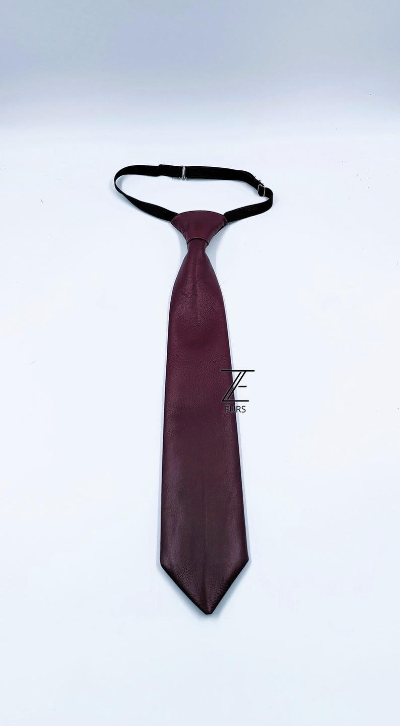 Peut inclure: Une cravate bordeaux fonc&eacute; avec une surface textur&eacute;e et une extr&eacute;mit&eacute; pointue. La cravate est dot&eacute;e d'une sangle de cou &eacute;lastique noire r&eacute;glable en haut. Un logo noir avec la lettre Z et le mot FURS est imprim&eacute; au centre du tissu de la cravate.