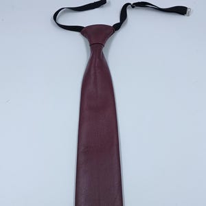 Peut inclure: Une cravate bordeaux fonc&eacute; fabriqu&eacute;e dans un mat&eacute;riau en cuir textur&eacute;. La cravate pr&eacute;sente une extr&eacute;mit&eacute; inf&eacute;rieure pointue et une structure de n&oelig;ud pr&eacute;-nou&eacute;. Elle comprend une sangle de cou &eacute;lastique noire r&eacute;glable avec une boucle coulissante en m&eacute;tal argent&eacute;.