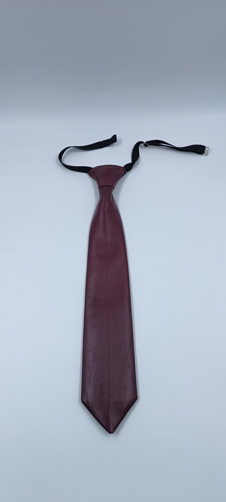 Peut inclure: Une cravate bordeaux fonc&eacute; fabriqu&eacute;e dans un mat&eacute;riau en cuir textur&eacute;. La cravate pr&eacute;sente une extr&eacute;mit&eacute; inf&eacute;rieure pointue et une structure de n&oelig;ud pr&eacute;-nou&eacute;. Elle comprend une sangle de cou &eacute;lastique noire r&eacute;glable avec une boucle coulissante en m&eacute;tal argent&eacute;.