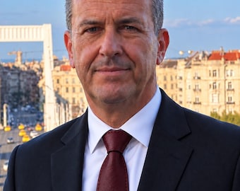 Peut inclure: Une personne d'âge moyen aux cheveux courts, foncés et texturés, portant une chemise blanche à col, une cravate bordeaux foncé et une veste de costume noire ajustée. La personne a une expression neutre devant un décor urbain.