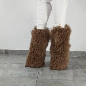 May include: A pair of brown, long-haired, curly faux fur boot covers worn over white pants. The textured, shaggy material extends from the ankle to the mid-calf, completely obscuring the footwear underneath, creating a voluminous, fuzzy appearance.