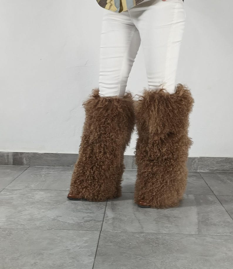 May include: A pair of brown, long-haired, curly faux fur boot covers worn over white pants. The textured, shaggy material extends from the ankle to the mid-calf, completely obscuring the footwear underneath, creating a voluminous, fuzzy appearance.