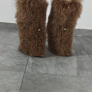 May include: A pair of tall, brown, shaggy faux fur boot covers worn over white pants. The textured, curly brown material extends from the mid-calf down to the base of the footwear, featuring a small gold-toned metal accent on the front of each.