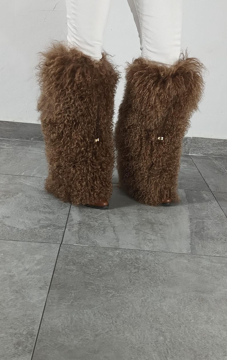 May include: A pair of tall, brown, shaggy faux fur boot covers worn over white pants. The textured, curly brown material extends from the mid-calf down to the base of the footwear, featuring a small gold-toned metal accent on the front of each.