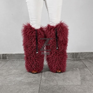 May include: A pair of burgundy, long-haired, curly fur leg warmers worn over white pants. The leg warmers feature black drawstring ties with silver-toned metal toggles. A black logo with the text ZE FURS is centered between the two leg warmers.