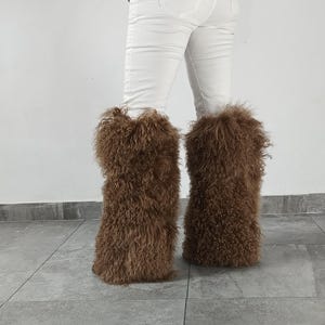 May include: A pair of brown, long-haired, shaggy faux fur leg warmers worn over white pants. The leg warmers feature a dense, curly texture that extends from the lower calf to the ankle, providing a voluminous appearance around the lower legs.